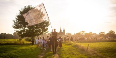 Łąka - Mężczyzna na szczudłach niosący flagę, za nim tłum ludzi. W tle drzewo i dwie kościelne wieże.