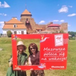 Dwie kobiety pozujące z dużych wymiarów pocztówką z napisem "Mazowsze poleca się na wakacje!". W tle Zamek w Liwie.