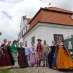 Osoby w historycznych strojach przed dworkiem barokowym.