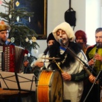 Grający i śpiewający muzycy w kolorowych strojach karnawałowych. w tle przystrojona choinka.