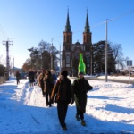 Tłom ludzi idących gęsięgo. Zimowa sceneria. W tle dwie wieże liwskiego kościoła.