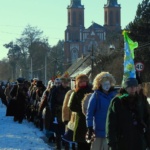 Tłom ludzi idących gęsięgo. Zimowa sceneria. W tle dwie wieże liwskiego kościoła.