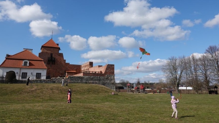 Dziecko puszczające latawiec. W tle przechodzący ludzie i Zamek w Liwie.