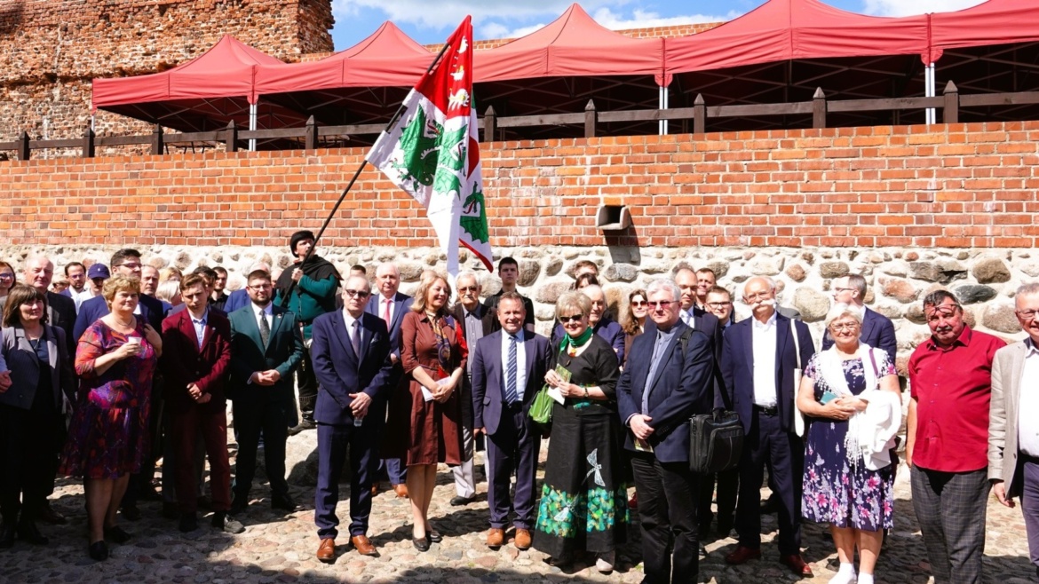 Grupa osób stoi przed czerwoną ścianą budynku, trzymając flagę oraz uśmiechając się do aparatu w słoneczny dzień