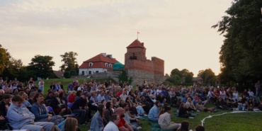 W tle zamek, na pierwszym planie grupa ludzi siedzących na krzesłach i kocach