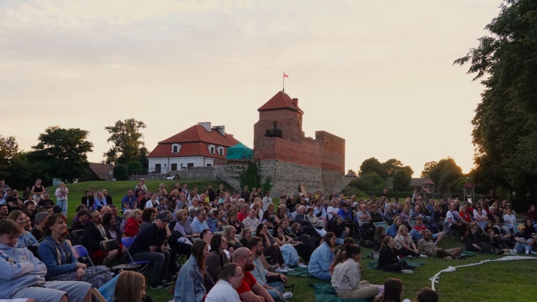 W tle zamek, na pierwszym planie grupa ludzi siedzących na krzesłach i kocach