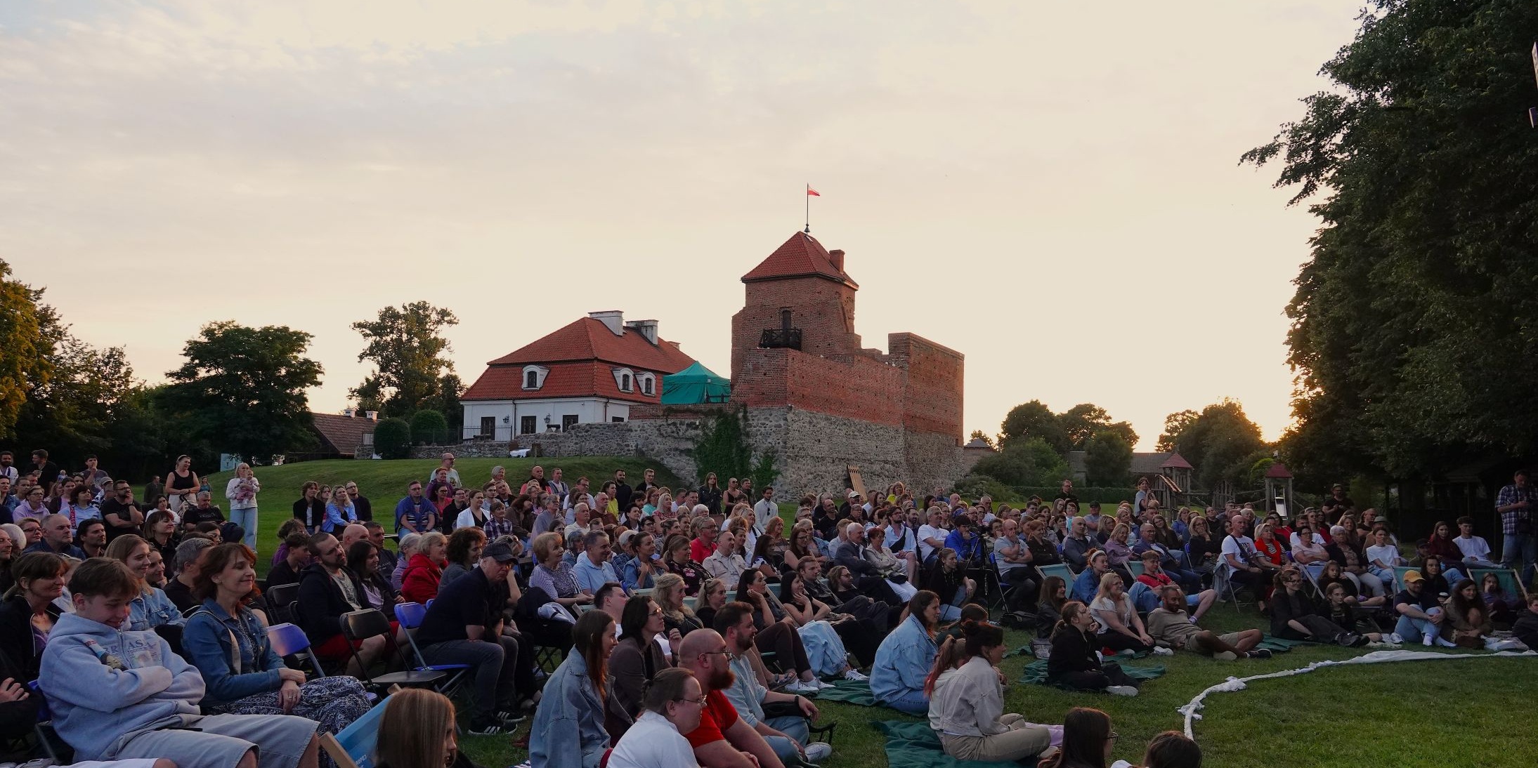 W tle zamek, na pierwszym planie grupa ludzi siedzących na krzesłach i kocach