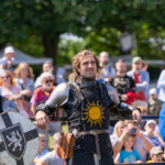 Trzech rycerzy w zbroi, jeden z nich z tarczą, a w tle tłum widzów na festiwalu kultury średniowiecznej
