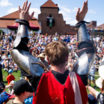 Mężczyzna w zbroi unosi ręce w geście powitania, a w tle widać tłum widzów zgromadzonych na wydarzeniu historycznym