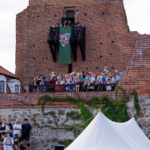 Liczna grupa ludzi zgromadzona na murze zamku, z flagą w tle, w słoneczny dzień