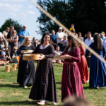 Kobiety w średniowiecznych strojach tańczą na festiwalu, trzymając misy, podczas gdy tłum widzów obserwuje z zainteresowaniem