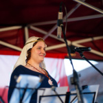 Kobieta w historycznym stroju z białym nakryciem głowy i biżuterią stoi przy mikrofonie, uśmiechając się w stronę publiczności