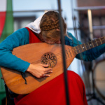 Muzyk z długimi włosami gra na lutni, pochylając się nad instrumentem, ubrany w kolorowy strój ludowy