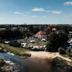 Teren festiwalu na tle rzeki, z licznymi uczestnikami, namiotami, parkingiem pełnym samochodów oraz zielonym krajobrazem