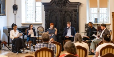 Sześć osób siedzących w rzędzie, czytających teksty, w eleganckim pomieszczeniu z historycznymi elementami wystroju